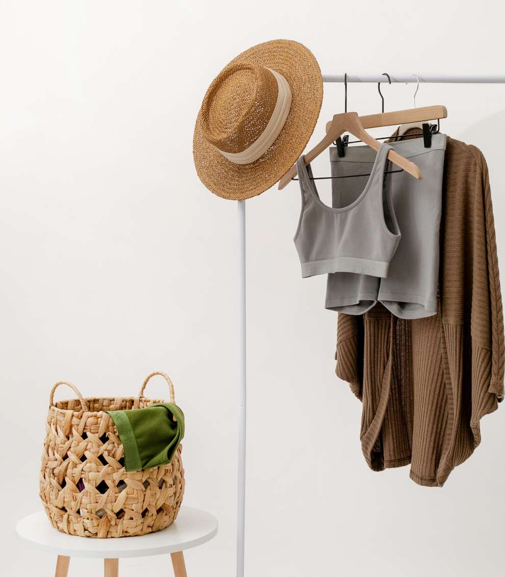 Photo of a minimalist wardroe with amrêve organic pima cotton clothing. There is a clothing rack with the amrêve organic bralette and bike shorts in grey and a brown cardigan. There is a straw hat resting on the clothing rack and a wicker basket.